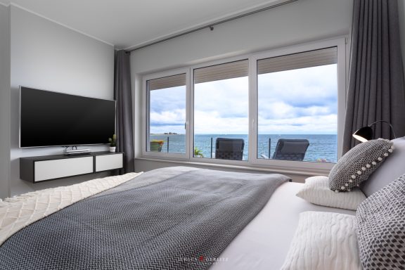 Kingsize Bett und TV vor Fenster zum Wasser und Leuchtturm - Villa Blue Horizon
